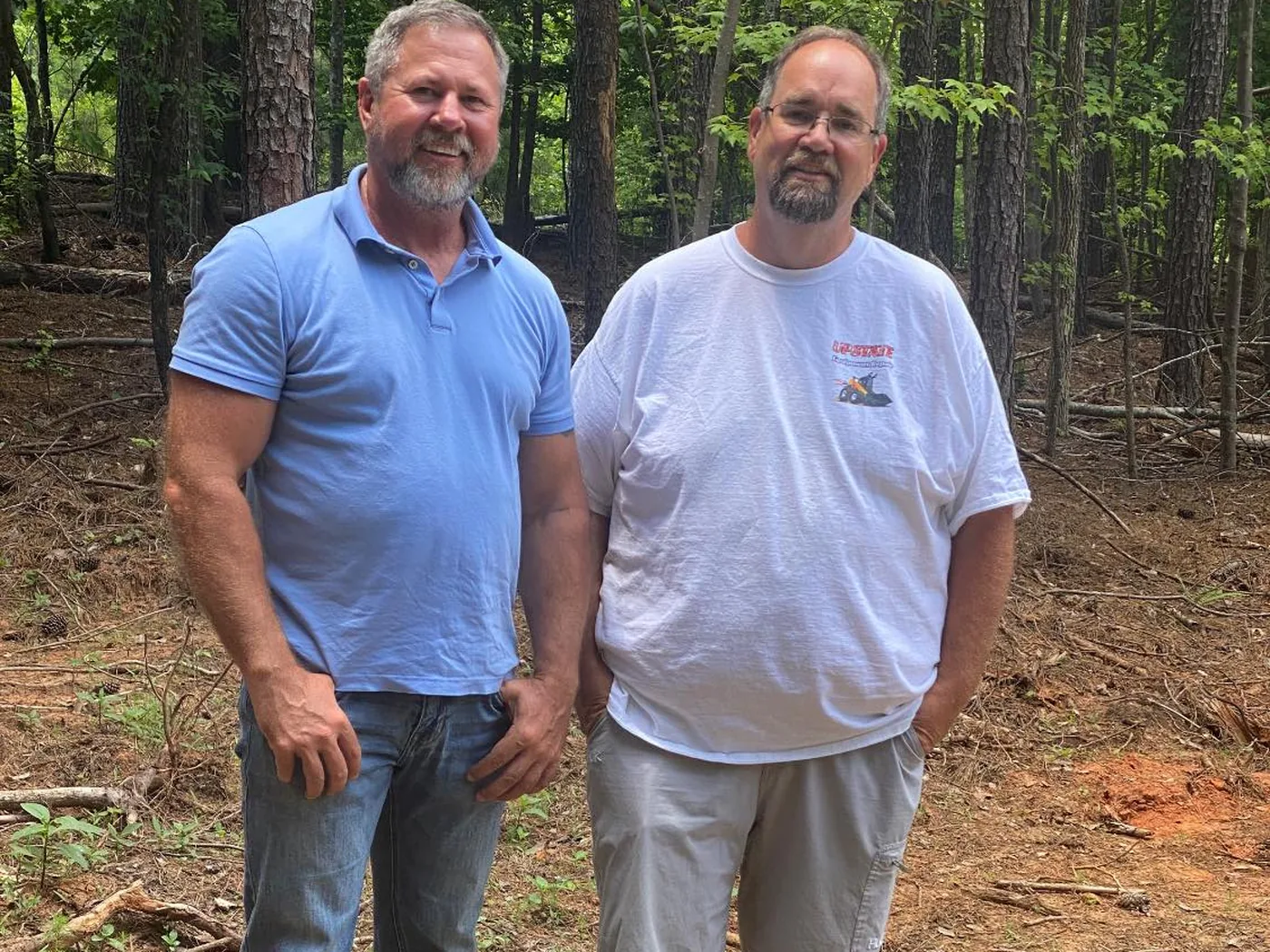 Two men standing side by side beside a newly finished white well casing on a wooded lot