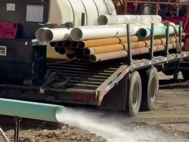 Water blowing out of a discharge hose across open ground beside a trailer of drill pipe and a water tank