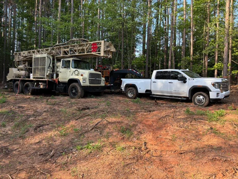 A drill rig and a white pickup parked on an open cleared tract of red clay ringed by pines