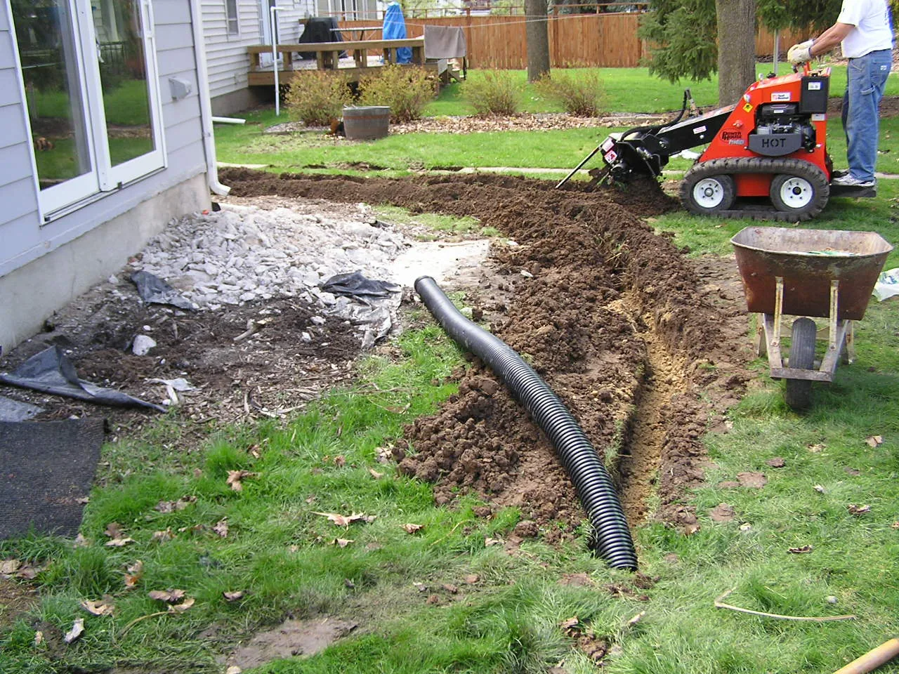A compact walk-behind trencher cutting a narrow trench across a lawn toward a house with pipe laid alongside
