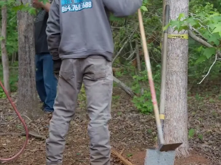 A crew member in a Core Well Drilling hoodie digging with a shovel beside the drill rig on a red clay site