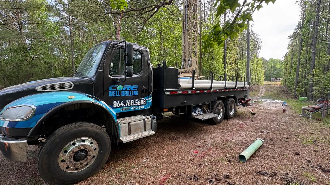 The wrapped Core Well Drilling flatbed truck parked on a wooded lane with well casing pipe on the ground