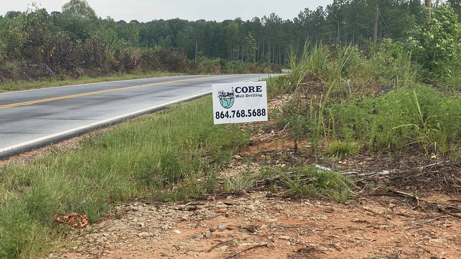 A Core Well Drilling yard sign at the edge of a cleared building lot along a two-lane country road