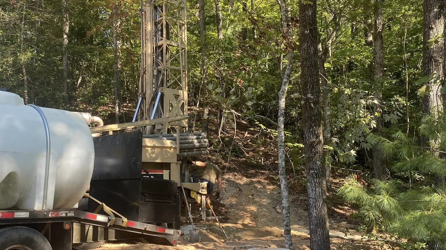A water tank truck and drill rig parked on a wooded job site with rock spoil in the foreground