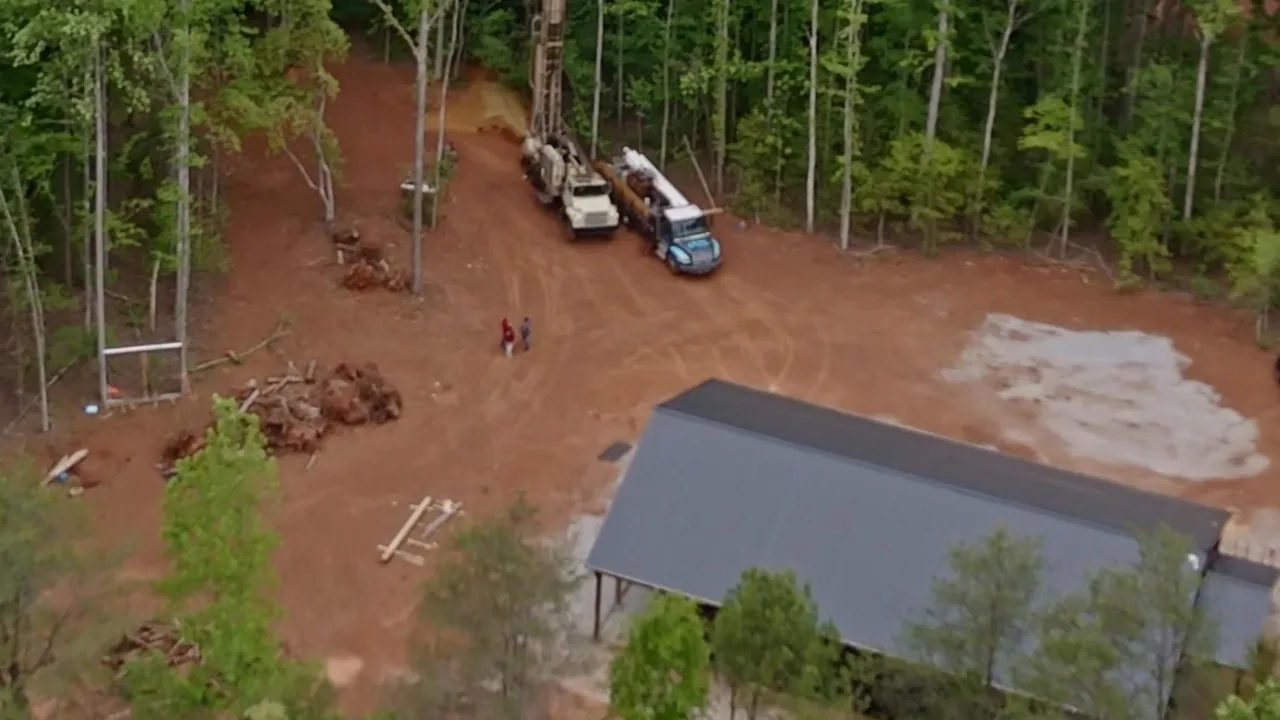 Aerial view of two work trucks on a cleared red clay lot in the woods, the kind of rural land we drill around Greenwood