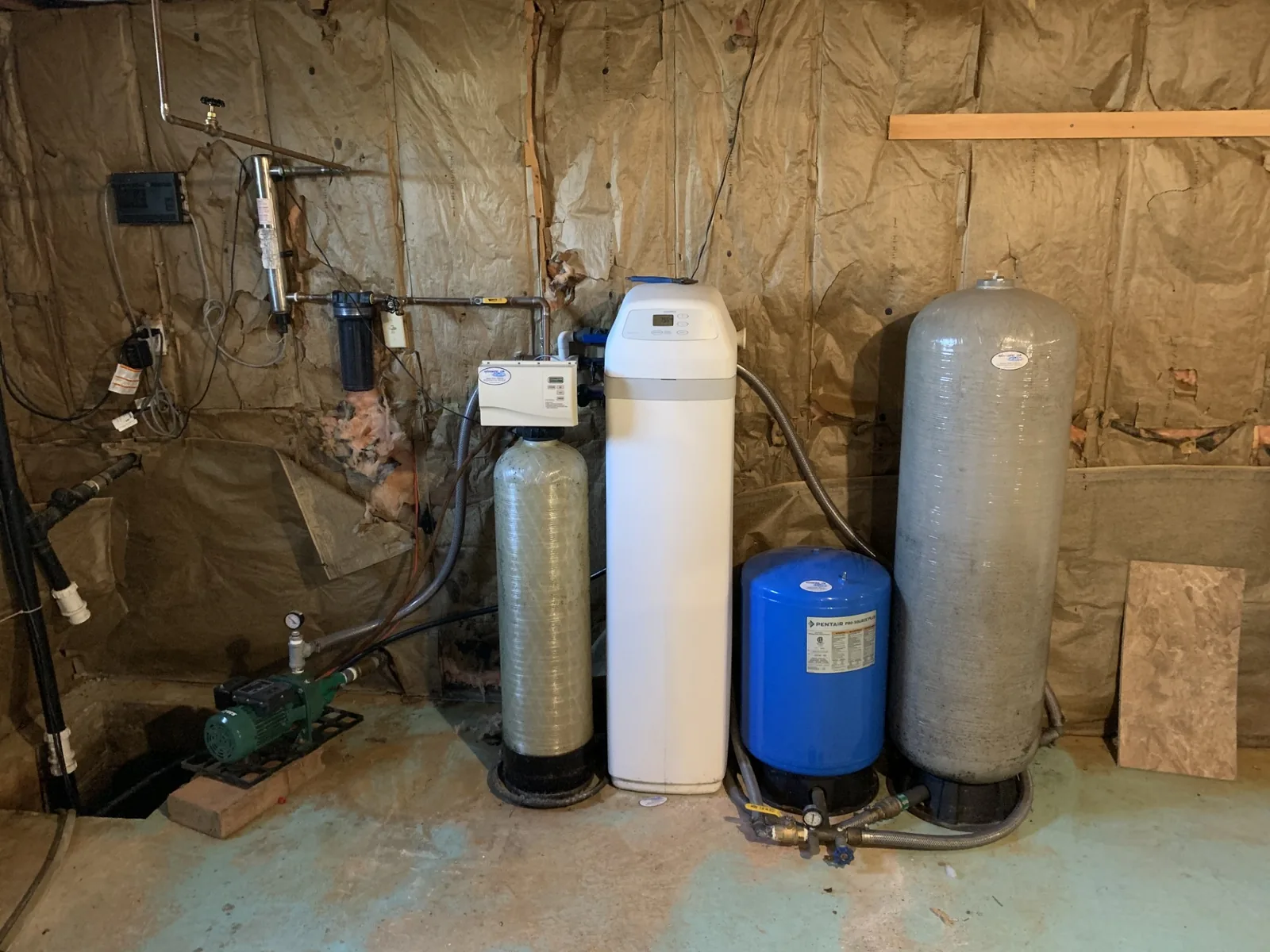 A basement well water setup with a jet pump, sediment filter, softener and a blue pressure tank against the wall
