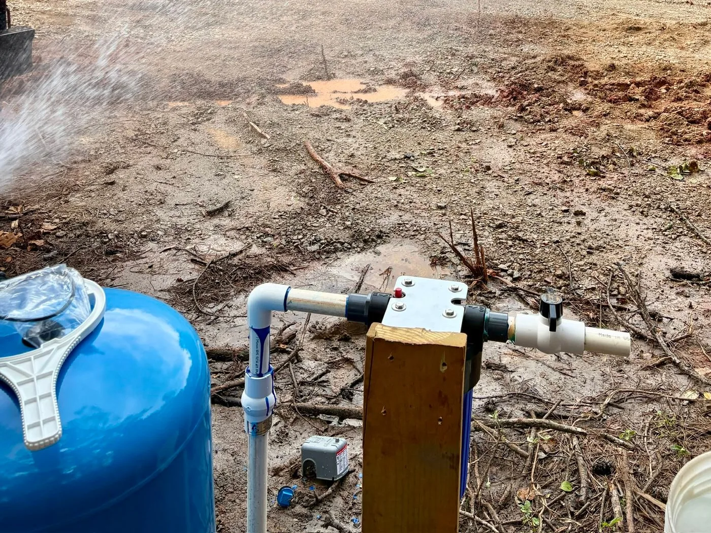 A blue pressure tank and pressure switch plumbed up at a well site while water blows from the new well