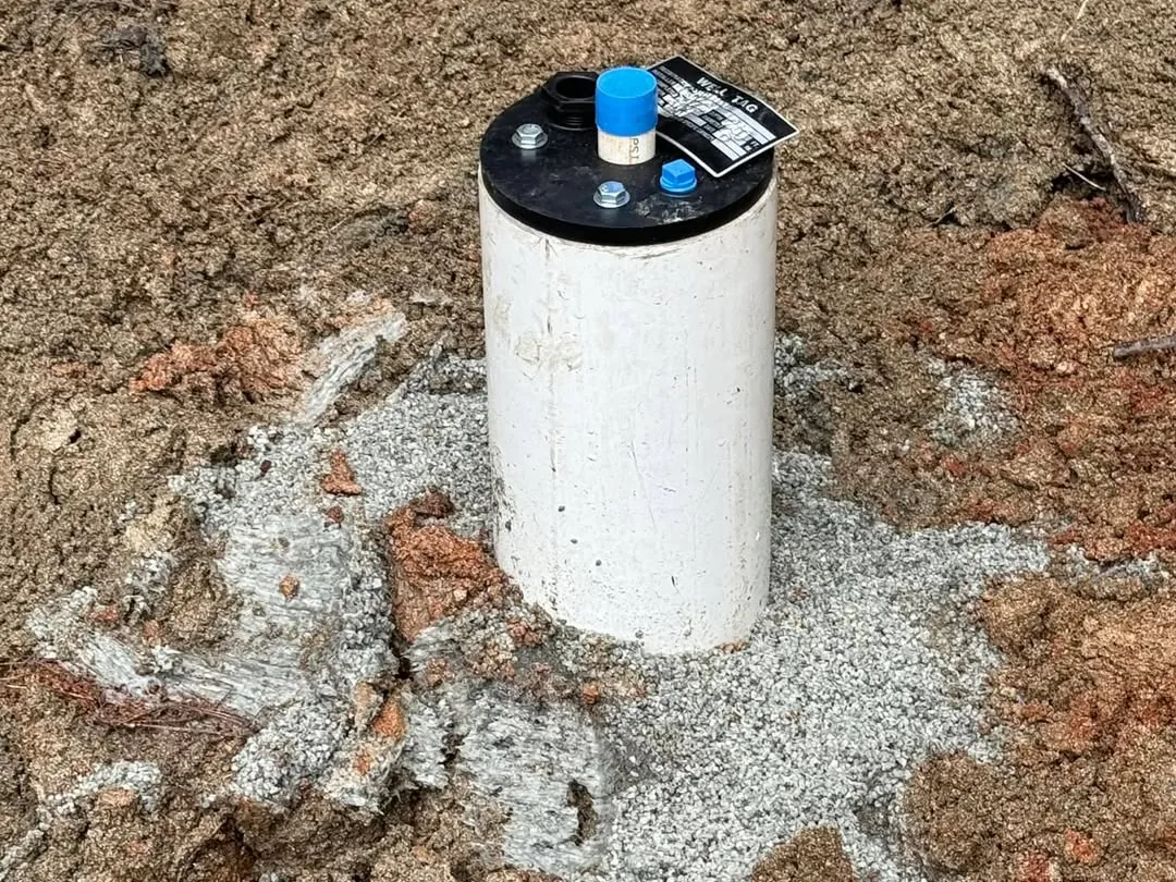 A finished white PVC well casing with a black sanitary well cap set in fresh grout and red clay