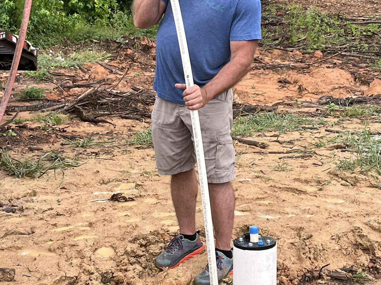 A worker lowering a length of white PVC pipe into a new well casing on a freshly cleared red clay lot