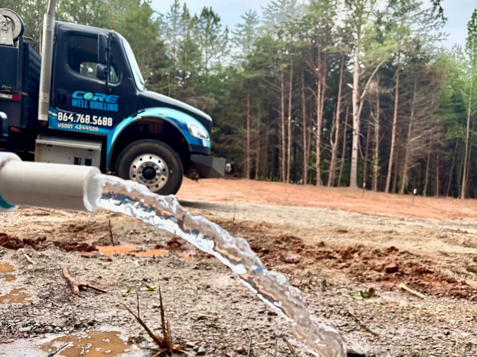 Clear water arcing out of a white discharge pipe onto red clay at a finished well, with a Core Well Drilling truck behind