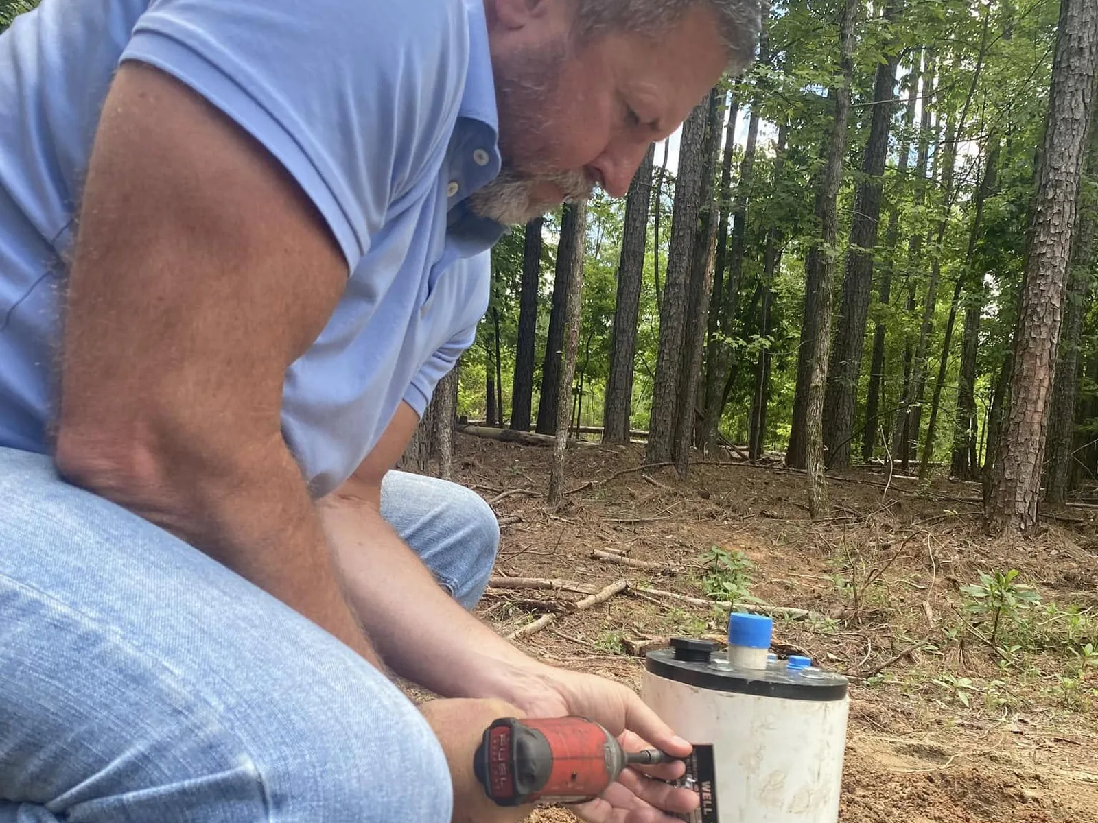 A well technician crouched at a white well casing, working on the black well cap with a cordless drill